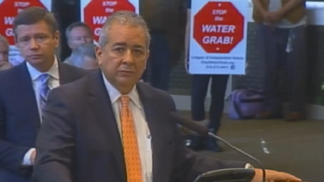 This screengrab shows SAWS CEO Robert Puente answering questions from City Council last October about the Vista Ridge pipeline. Protestors stand behind him.