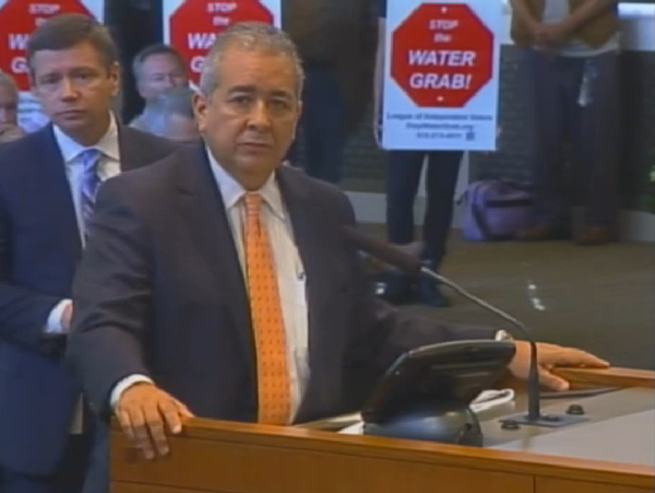 This screen grab of a City Council meeting shows San Antonio Water System President and CEO Robert Puente answering questions as protestors stand behind him. - San Antonio Current