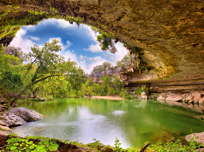 Water falls off the top section of rock into Hamilton Pool on a sunny day. - Shutterstock / dibrova