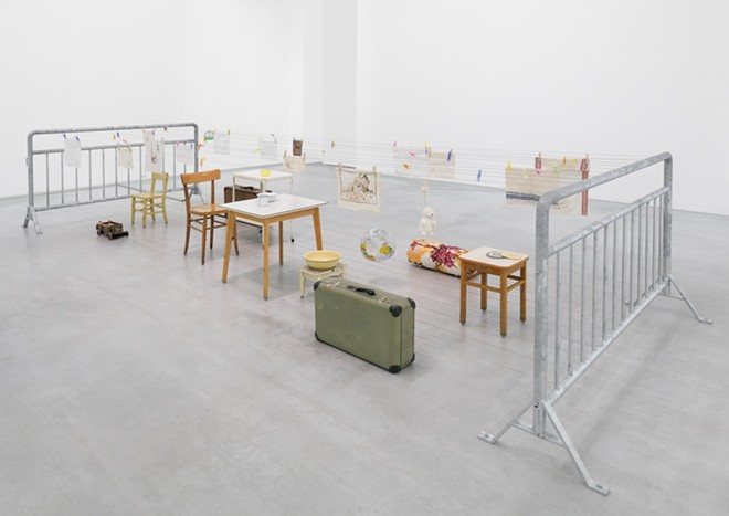 Mona Hatoum (b. 1952, Beirut, Lebanon; lives London, UK). Mobile Home II, 2006. Furniture, household objects, suitcases, galvanized steel barriers, three electric motors, and pulley system. 46 7/8 x 86 5/8 x 236 1/4 in. - 2023.2. - Mona Hatoum, courtesy of Ruby City